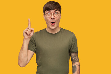 I have an idea! Young attractive handsome male, wears green t-shirt, round glasses, point with a finger upwards, opened eyes widely, with amazed facial expression. Isolated over yellow background.