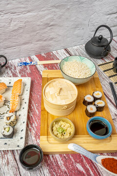 Image of vintage red paint table with plates of asian food and sushi. Steam dim sum basket, soy sauce, rice bowl, traditional teapot, chopsticks, ginger and wasabi, salmon makis.
