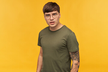 Portrait of young male, looks directly into camera with angry, negative facial expression. Wears green t-shirt and round glasses. Isolated over yellow background.