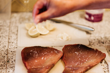 Zubereitung von mediterranem Thunfisch Steak, Filet. Eine Hand streut Salz über den Fisch, das Steak. Im Hintergrund liegen Zitronen.