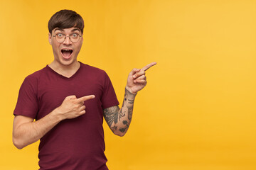 Indoor shot of young male, wears red t-shirt and round glasses, indicates with both hands at copy space, while smiling. Isolated over yellow background.