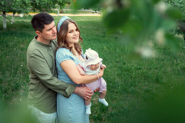 Blonde mother with long loose hair lulls baby daughter and talks to smiling husband in green shirt standing in park with blooming trees, copyspace