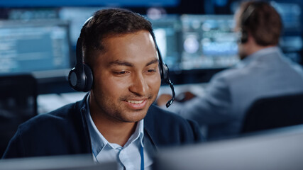 Close Up Portrait of a Joyful Technical Customer Support Specialist Talking on a Headset while Working on a Computer in a Call Center Control Room Filled with Computer Display Screens and Data Servers