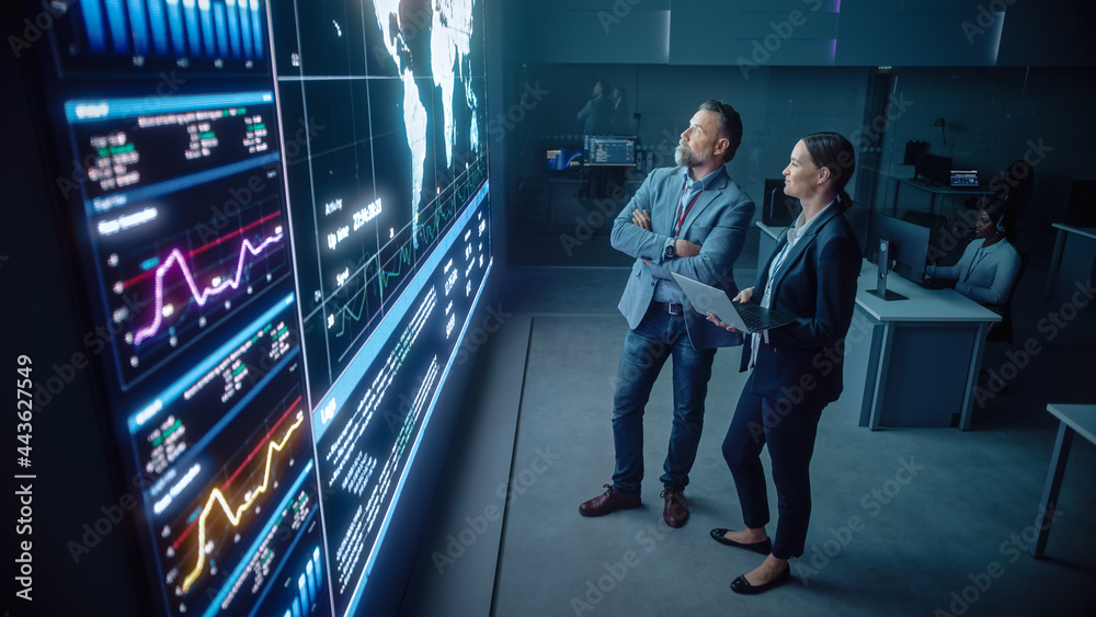 Fototapete Project Manager and Computer Science Engineer Talk while Using Big Screen Display and a Laptop, Showing Infrastructure Infographics Data. Telecommunications Company System Control and Monitoring Room.