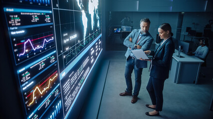 Project Manager and Computer Science Engineer Talk while Using Big Screen Display and a Laptop, Showing Infrastructure Infographics Data. Telecommunications Company System Control and Monitoring Room.