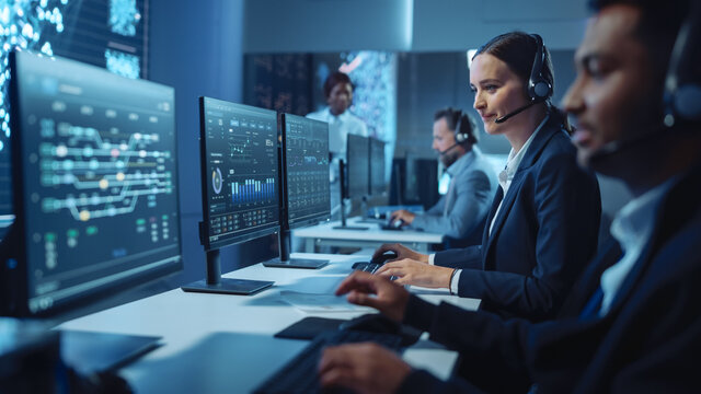 Happy Confident Technical Customer Support Specialist Having A Headset Call While Working On A Computer In A Dark Monitoring And Control Room Filled With Colleagues And Display Screens.