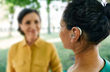 Adult woman with a hearing impairment uses a hearing aid to communicate with her female friend at city park. Hearing solutions - 443628321