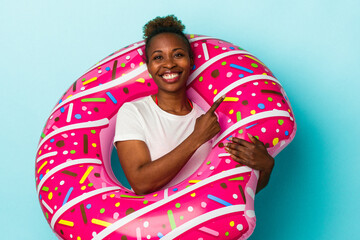 Young african american woman with inflatable donut isolated on blue background smiling and pointing aside, showing something at blank space.