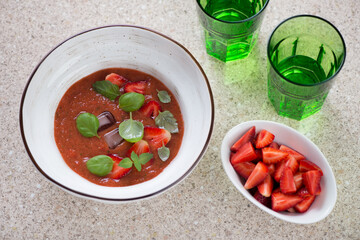 Bowl of cold gazpacho made of red tomatoes and fresh strawberry, elevated view on a beige marble background, horizontal shot