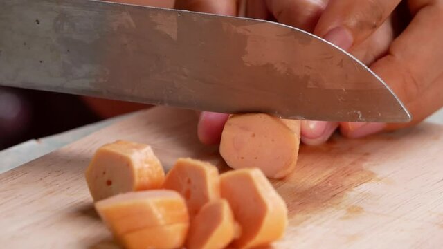 Close up of a knife cuts the sausage into small pieces. Mother holds her daughter's hand to teach her how to cut sausages to prepare them for making Borona sauce over spaghetti at home.