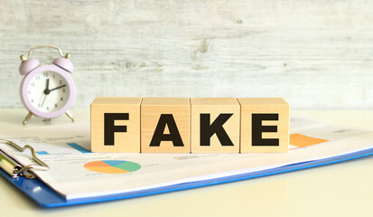 Wooden cubes lie on a folder with financial charts on a gray background. The cubes make up the word FAKE.