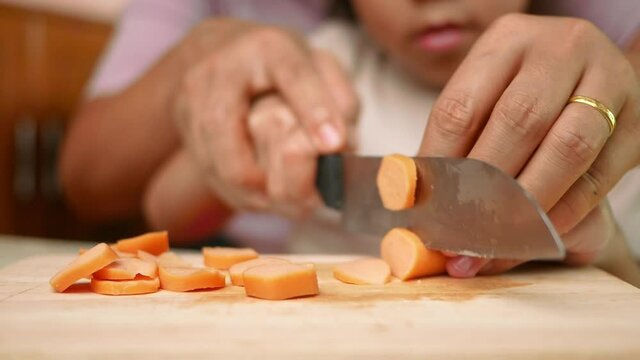 Close up of a knife cuts the sausage into small pieces. Mother holds her daughter's hand to teach her how to cut sausages to prepare them for making Borona sauce over spaghetti at home.