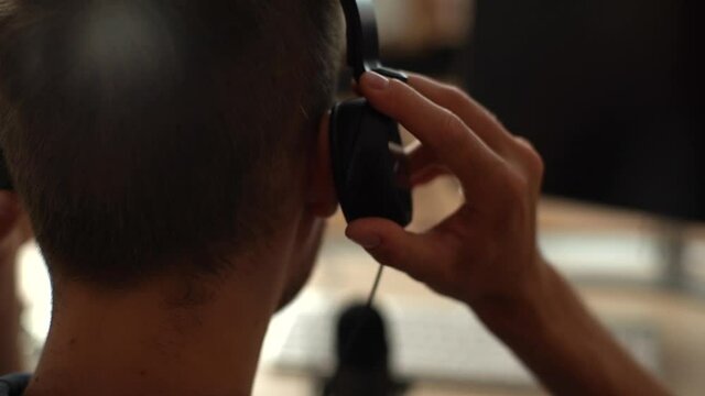 Close-up back view of unrecognizable blogger male putting headphones on head sitting at desk with microphone and computer monitor. Closeup rear view of young man puts headphones on head indoors.