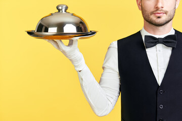 Handsome elegant waiter holding tray and cloche ready to serve on yellow background