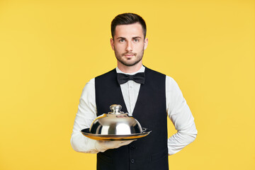 Handsome elegant waiter holding tray and cloche ready to serve on yellow background