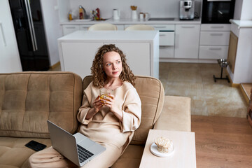 Beautiful young European woman in casual clothing using laptop and smiling while relaxing in living room