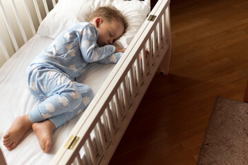 top view Cute little 2-3 years preschool baby boy kid sleeping sweetly in white crib during lunch rest time in blue pajama with pillow at home. Childhood, leisure, comfort, medicine, health concept