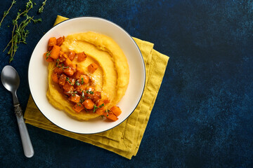 Polenta with butter, pumpkin, garlic, savory and parmesan cheese in white bowl on dark blue concrete background. Traditional Basic Italian food, vegan food. Top view. Copy space.