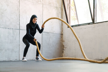 Young muslim woman in hijab working out with battle ropes indoors