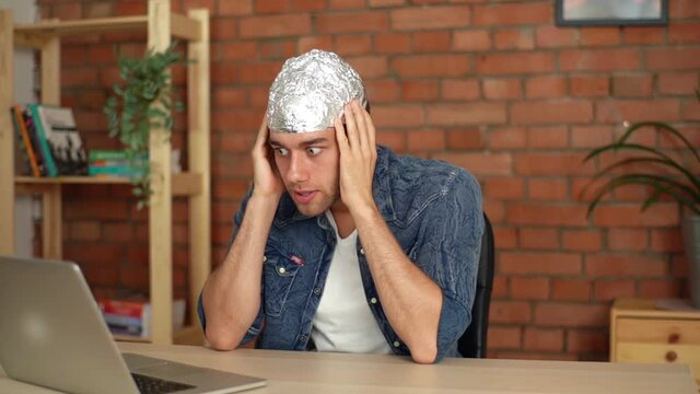 Close-up of scared conspiracy theorist wearing foil hat watching online video using laptop sitting at home. Shocked paranoid young man search in internet information about conspiracy and watching film