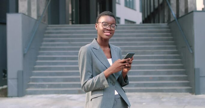 Attractive, adult multiracial woman in chic formal wear standing near the business center. She looking at camera and holding telephone with mail email in hands