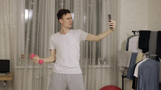 Funny man doing fitness exercise with dumbbell and taking selfie on smartphone while having fun during crazy sport training at home 