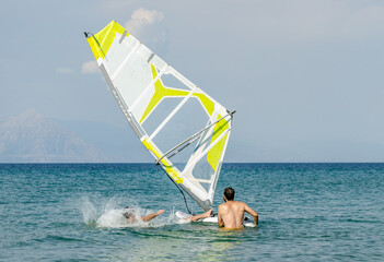 Naklejka premium Man Fell from a Windsurfing Board in the Sea. Summer Water Sport Activities in Greece. Horizon Line in Background. Surf Teacher is Watching and Helping