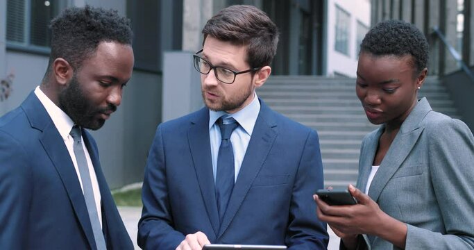 Portrait of outgoing young male showing information on tablet to his colleagues. They standing at the street and chatting. Conference concept