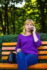 Defocus serious caucasian blond woman talking, speaking on the phone outside, outdoor. 40s years old woman in purple blouse in park. Adult person using phone. Vertical. Out of focus