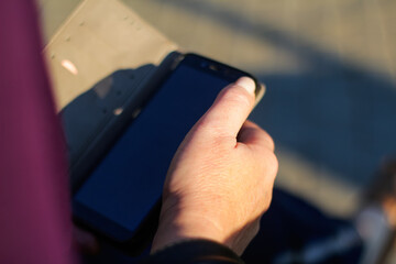 Defocus close-up female hands searching on the phone outside, outdoor. Adult woman in purple blouse using smartphone while sitting on a bench in the park. Copy space. Out of focus