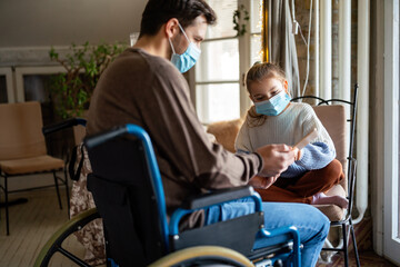 Father with disability in wheelchair using tablet at home with child while wearing masks.