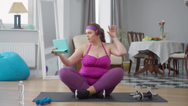 Confident plus-size Caucasian woman enjoying music in headphones sitting on exercise mat filming video blog. Obese young lady blogging training at home indoors. Confidence and self improvement