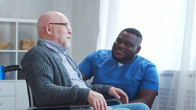 African-American caregiver and old disabled man in a wheelchair. Professional nurse and handicapped patient in a nursing home. Assistance, rehabilitation and health care.