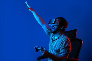 Young bearded man in VR glasses screams and celebrates winning in virtual game, sitting on chair...