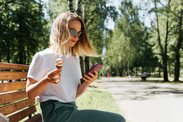 Young woman relaxing on a park bench and using a mobile phone. Nice girl eating ice cream and...