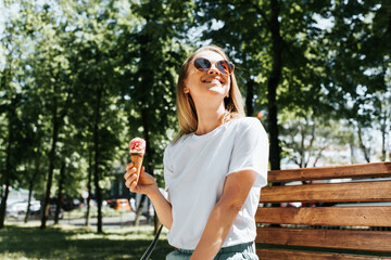 Joyful young woman relaxing on a park bench looking up enjoying the sun and summer weather. Cute smiling girl eating ice cream in a waffle cone, outdoors. Summer lifestyle - obrazy, fototapety, plakaty