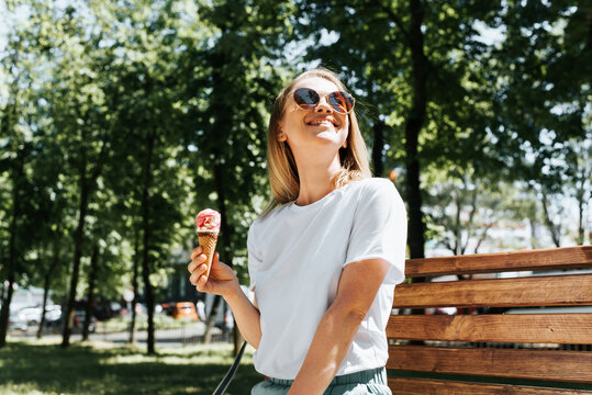 Joyful young woman relaxing on a park bench looking up enjoying the sun and summer weather. Cute smiling girl eating ice cream in a waffle cone, outdoors. Summer lifestyle