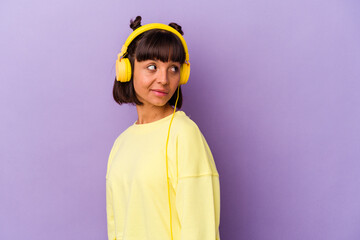 Young mixed race woman listening to music isolated on purple background looks aside smiling, cheerful and pleasant.