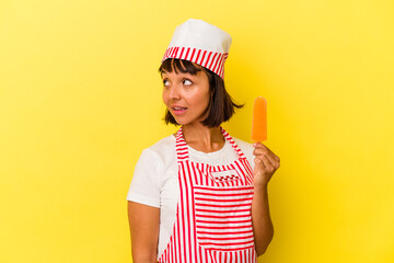 Young mixed race ice cream maker woman holding an ice cream isolated on yellow background looks aside smiling, cheerful and pleasant.