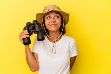 Young mixed race explorer woman holding binoculars isolated on yellow background dreaming of...