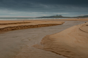 Dunstanburgh Castle in Northumberland, England, taken with the river meandering in the foreground & with cloudy skies overhead