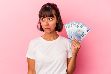 Young mixed race woman holding bills isolated on pink background confused, feels doubtful and unsure. - obrazy, fototapety, plakaty