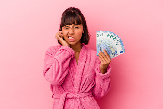 Young Mixed Race Woman Wearing A Bathrobe Holding Bills Isolated On Pink Background Covering Ears With Hands.