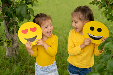 Two little funny girls are holding smile faces with different emotions in their hands. Children are having fun in the park. World emoji Day. Anthropomorphic smiley face.  - obrazy, fototapety, plakaty