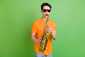 Naklejka premium Photo of happy positive young man wear sunglass hold hands play saxophone isolated on green color background
