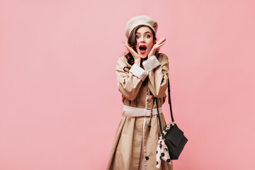 Lady with red lips is looking at camera in surprise. Woman in stylish trench coat posing with crossbody bag on pink background