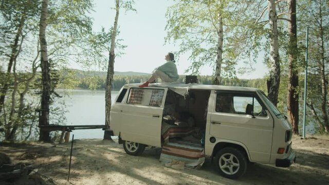 Zoom in wide shot of woman sitting on roof of camper van and enjoying view of scenic nature and lake