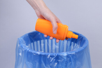 Hand throws plastic sunblock bottle into trash bin with package on gray background