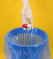 Wooden hand throws pill into trash bin with package on yellow background