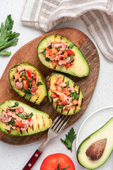 Grilled avocado stuffed with tuna, tomatoes, parsley, and olive oil on a cutting board. Top view, grey background